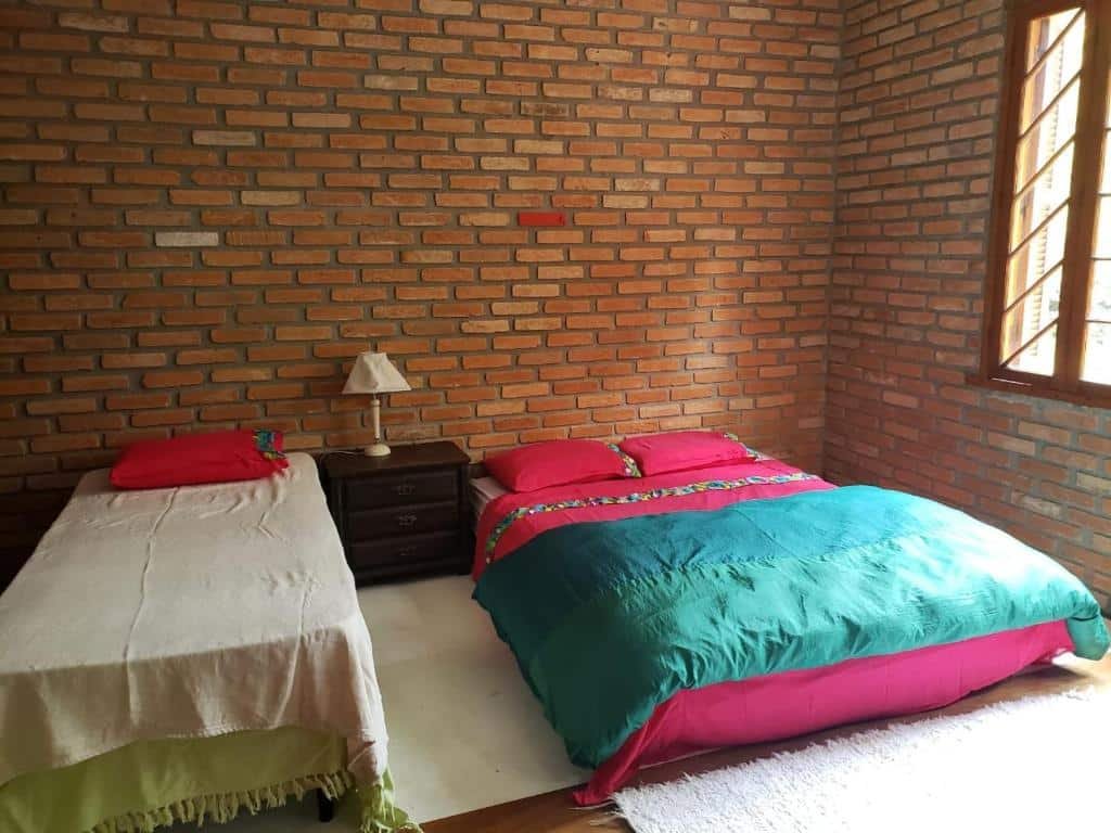 Quarto na Toca da Capuava. Uma cama de solteiro no canto esquerdo, uma cômoda no meio e uma cama de casal no lado direito. Uma janela no canto direito.