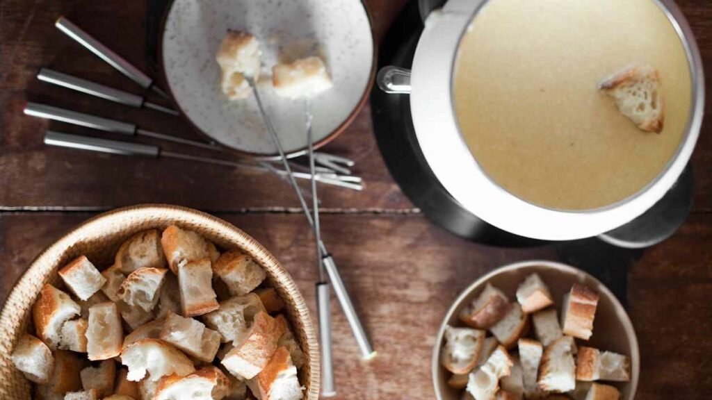 Fondue de queijo em uma mesa com tigelas de cubos de pão e garfos de fondue. Um cubo de pão é mergulhado na panela de fondue. Pertencem ao restaurante Crytal em Jungfrau.