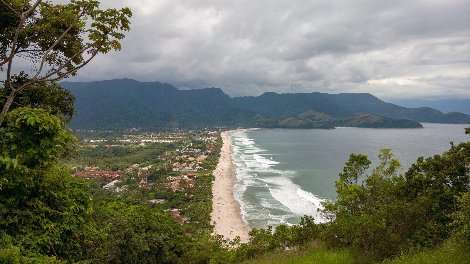 A imagem mostra uma vista panorâmica de Maresias, uma famosa praia localizada em São Sebastião, no estado de São Paulo, Brasil. A partir de um ponto elevado, pode-se ver a extensão da praia de Maresias, com seu longo trecho de areia branca margeado pelo oceano azul. As ondas quebram suavemente ao longo da orla. Ao fundo, a imagem exibe a vegetação exuberante e montanhosa que caracteriza a região, com montanhas verdes que se elevam majestosas. A vegetação densa, típica da Mata Atlântica, cerca tanto a praia quanto a área urbana. Na parte inferior da imagem, há um vislumbre das construções e casas que compõem a comunidade de Maresias, rodeadas por uma rica vegetação. O céu está coberto por nuvens cinzentas, sugerindo um dia nublado, mas ainda assim realçando a beleza natural da paisagem costeira.