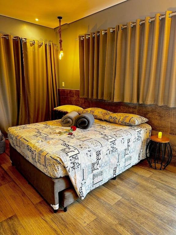 Foto de um quarto na pousada Águas Chalés Temporada que está representando o post sobre pousadas em Águas de São Pedro. Possui uma cama box com duas toalhas de banho e uma rosa em cima. Uma mesa de cabeceira está ao seu lado com uma vela. Há uma janela acima da cama e do seu lado esquerdo.