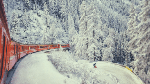 Um trem vermelho se move pelos Alpes Suíços por uma floresta coberta de neve, passando por um esquiador em uma encosta. A cena é envolta em cenário de inverno. Representa passeios na Suíça.