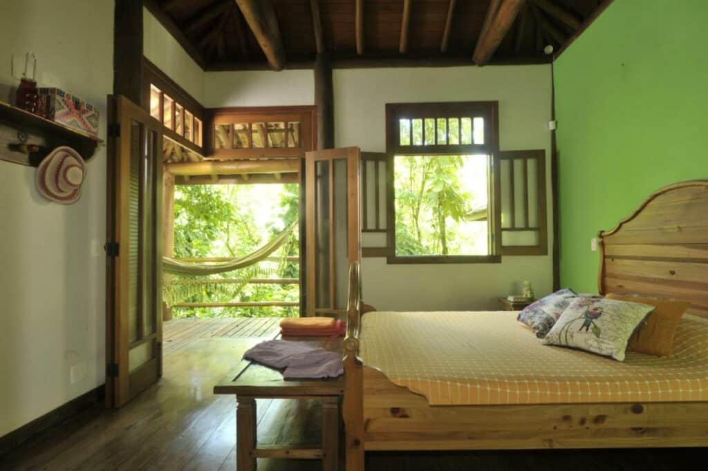 Quarto da Casa Sertão de Camburi com cama de casal do lado direto da imagem no pé da cama uma banco de madeira. Representa airbnb em São Sebastião.