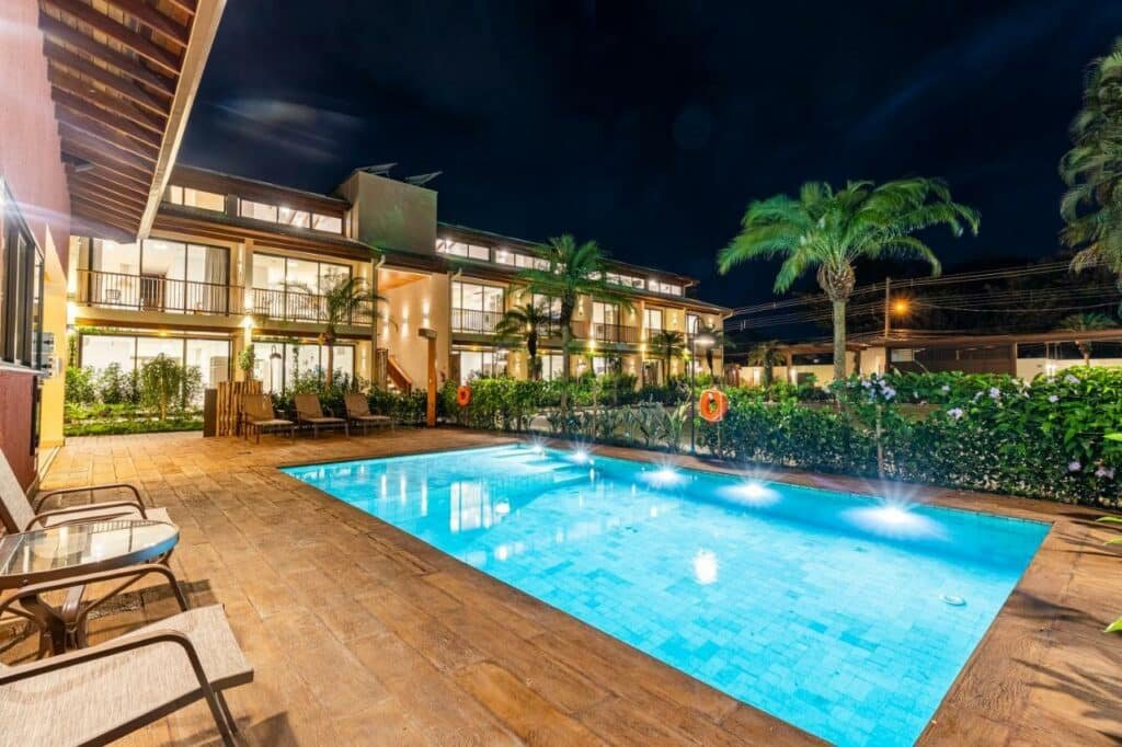Piscina do condomínio do airbnb "Anoa Maresias Studios". Ela é retangular, possui luzes noturnas e está em uma área com chão de madeira. Duas cadeiras e uma mesa estão por perto. Ao redor estão os apartamentos e algumas plantas.
