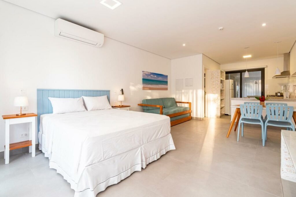 Quarto do Anoa Maresias Studios . Em primeiro plano está uma cama de casal com uma mesinha em cada lado. Perto dela e também encostado na parede está um sofá, de frente para uma mesa de quatro lugares. Ao fundo está a cozinha. Representa airbnb em São Sebastião.