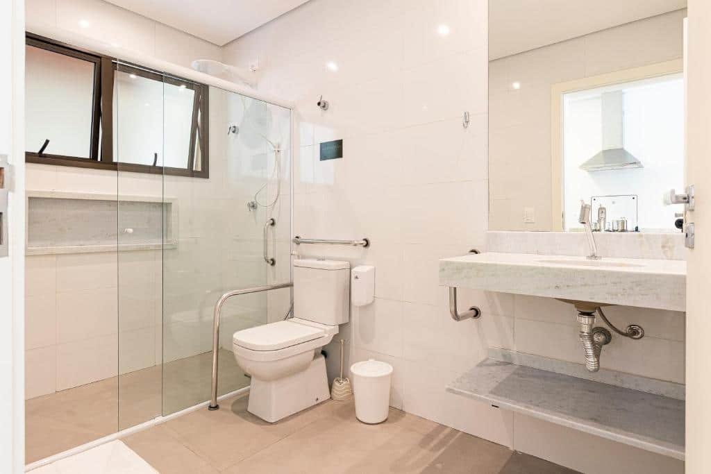 Banheiro acessível do airbnb Anoa Maresias Studios. Do lado esquerdo está o box, com uma barra de abaixo embaixo do chuveiro. Depois dele está o vaso, contornado por duas barras de apoio maiores. Já do lado direito há uma pia com espelho e uma pequena barra de apoio ao seu lado.