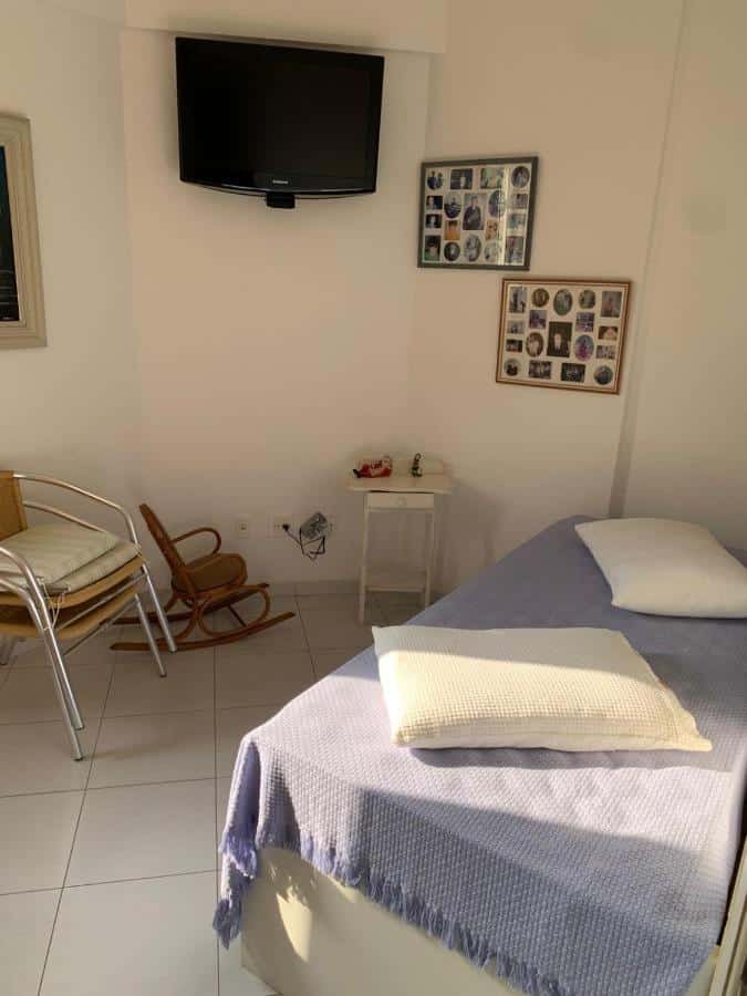 Quarto do Apartamento em Caraguatatuba em Frente a Praia, um dos airbnb em Caraguatatuba. Uma cama de solteiro com travesseiros e manta está encostada na parede ao lado direito, e ao seu lado esquerdo ficam duas cadeiras, uma mesinha de cabeceira e uma cadeira de balanço para crianças.