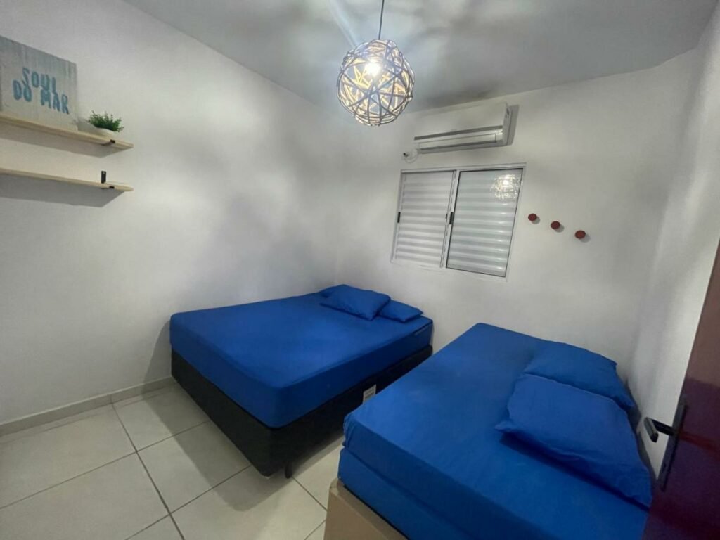 Quarto do Apê 06 Maresias. Duas camas estão lado a lado. Acima delas há uma janela e um ar-condicionado. No canto da imagem é possível ver duas prateleiras na parede.