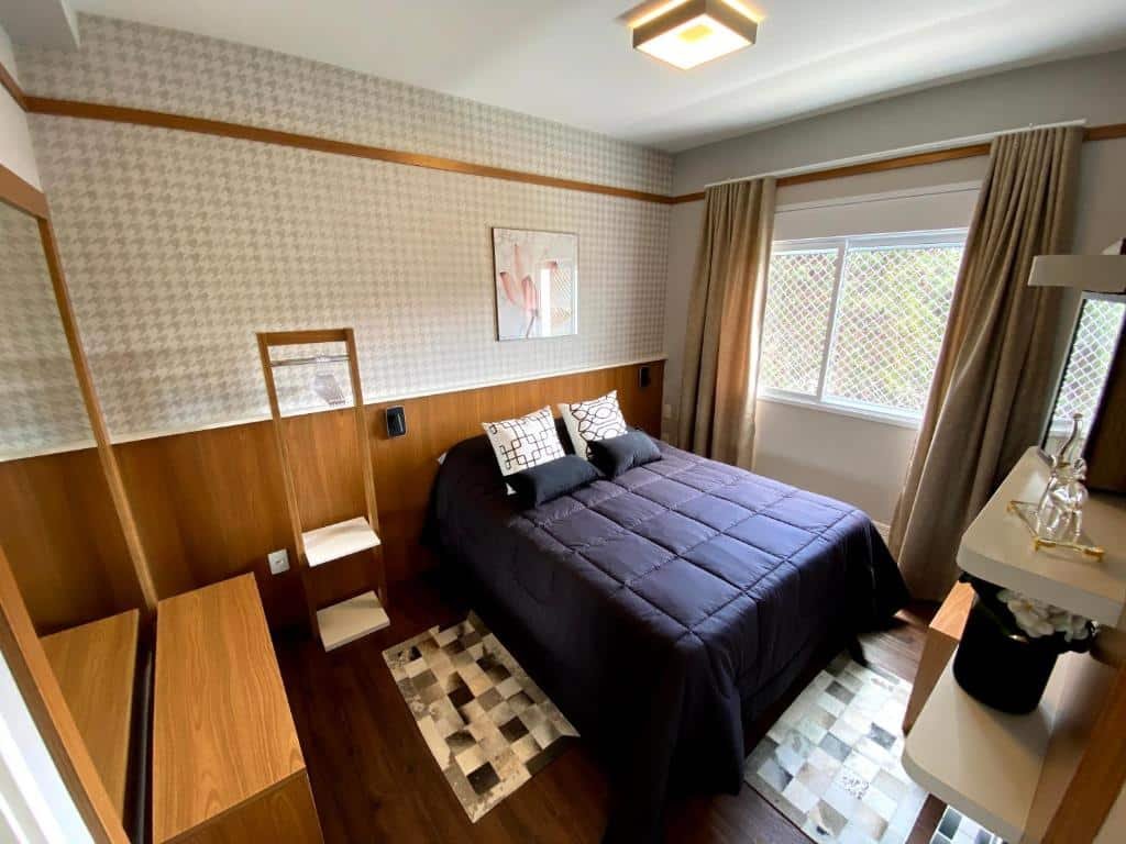 Quarto do Apartamento em Campos do Jordão ao lado do Capivari com uma cama de casal, janela com cortinas, estantes com enfeites, chão de madeira com tapetes, espelho e móveis com gavetas, para representar onde ficar em Campos do Jordão