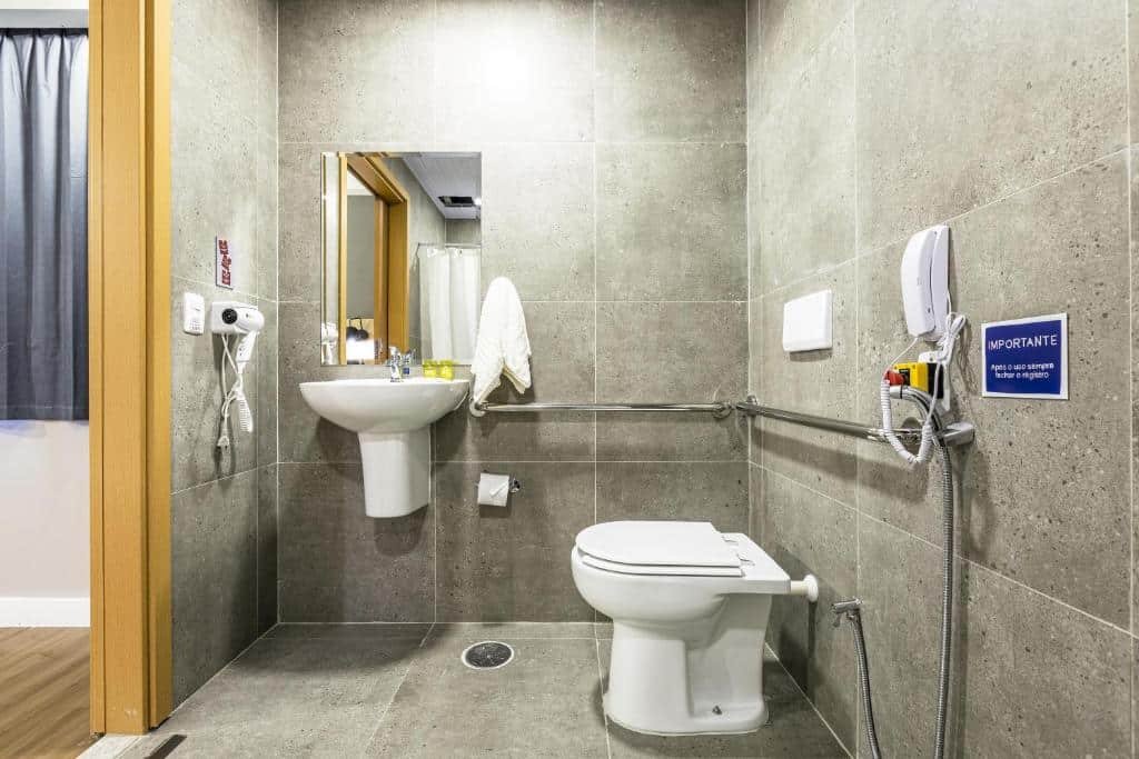 Banheiro com acessibilidade do 
Graal Inn Mairiporã. A porta de entrada no lado esquerdo, um secador e a pia com espelho. No lado direito o vaso sanitário com barras de apoio na parede e um mini chuveiro.