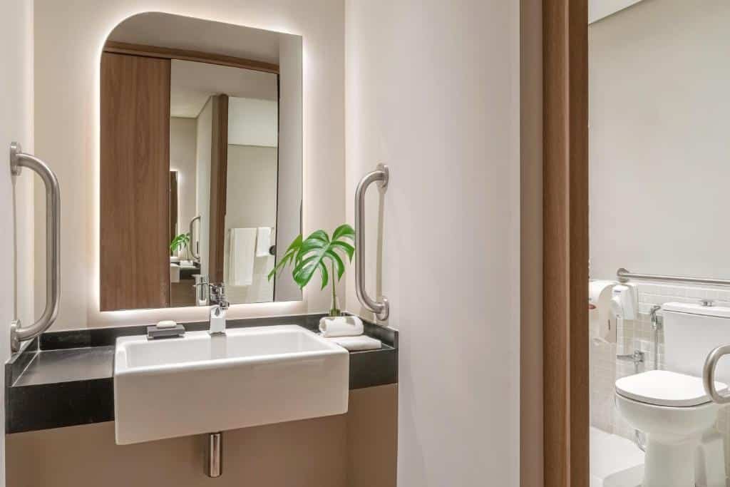 Banheiro acessível na Almenat Embu Das Artes Sao Paulo, Tapestry Collection. Uma pia com barras e espelho. Visão da porta com uma privada e barras de apoio.