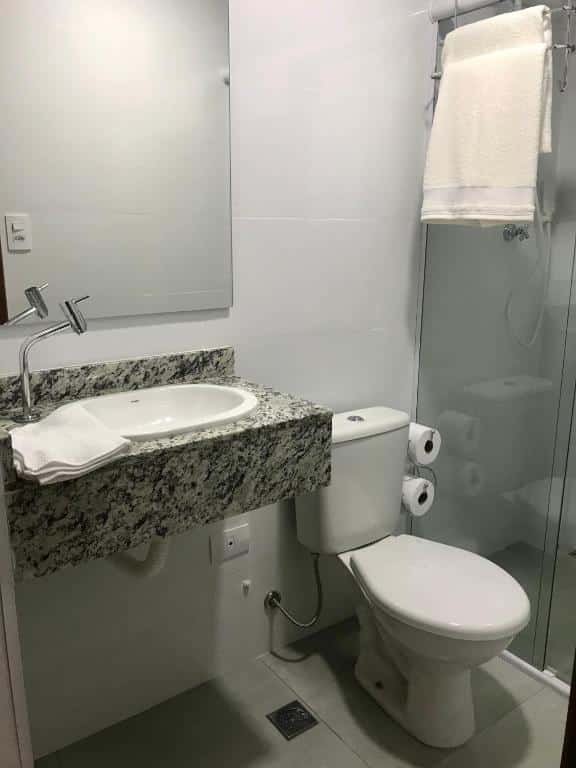 Imagem de um banheiro na Pousada Vila Barboza. Há um espelho, uma pia pequena e um vaso sanitário ao seu lado. Podemos ter uma visão parcial do box.