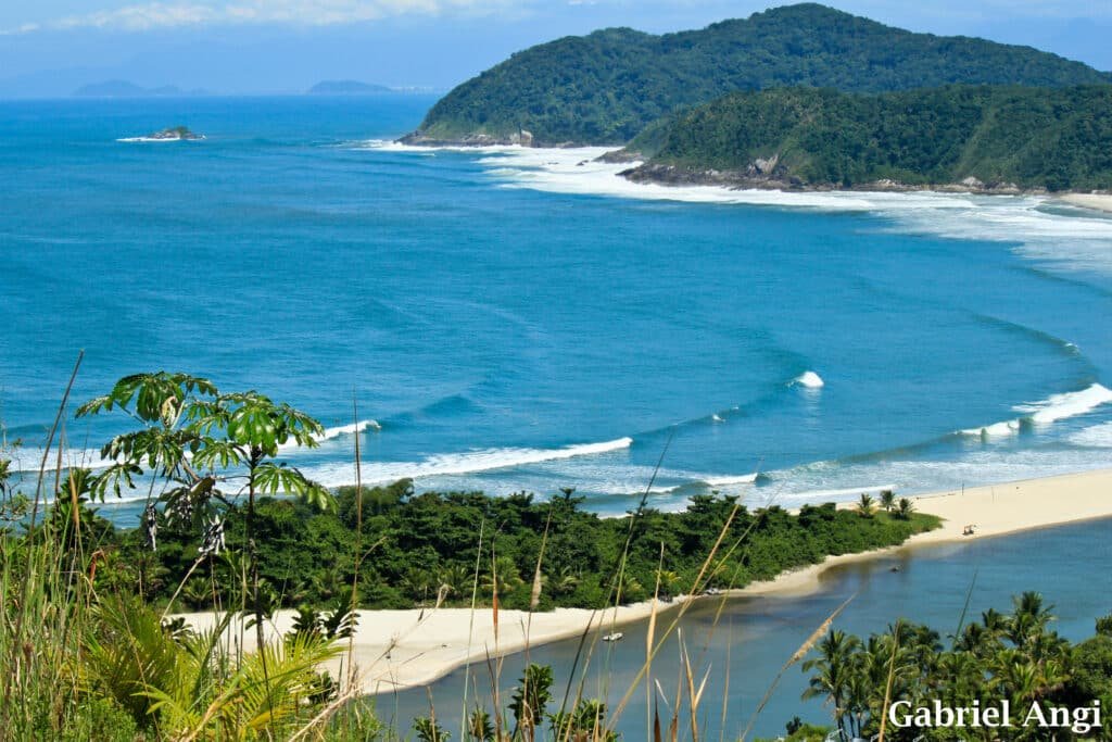 Vista aérea da Barra do Una com colinas verdejantes, praias arenosas e ondas azuis do oceano sob um céu limpo. É possível identificar o mar do lado esquerdo e o Rio Una no lado direito.