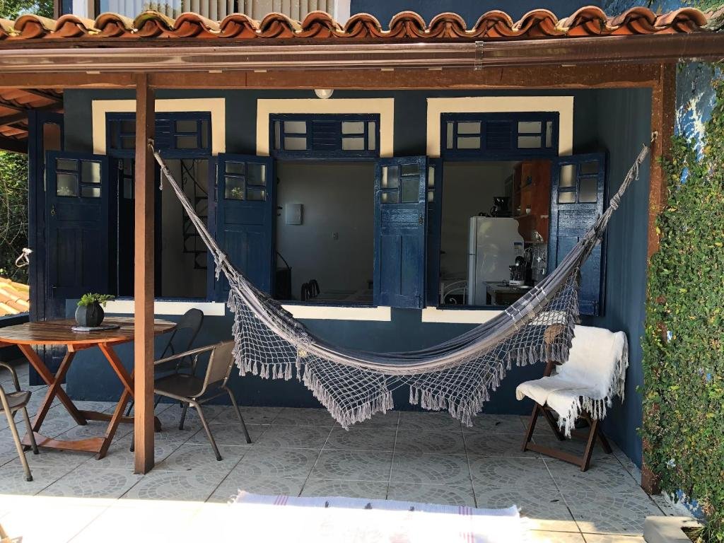 Área externa do airbnb Bergamota. Na varanda do airbnb há uma mesa redonda com cadeiras no lado esquerdo, no lado direito há uma rede de descanso e atrás está o airbnb.
