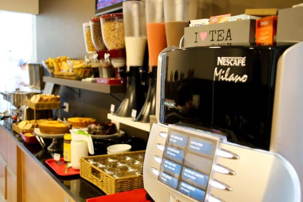 Mesa do café da manhã. Uma máquina de café na frente, atrás sucos e depois pães, bolachas e bolos. Na Pousada Du Valle. Foto para ilustrar post sobre Pousadas em Embu das Artes.