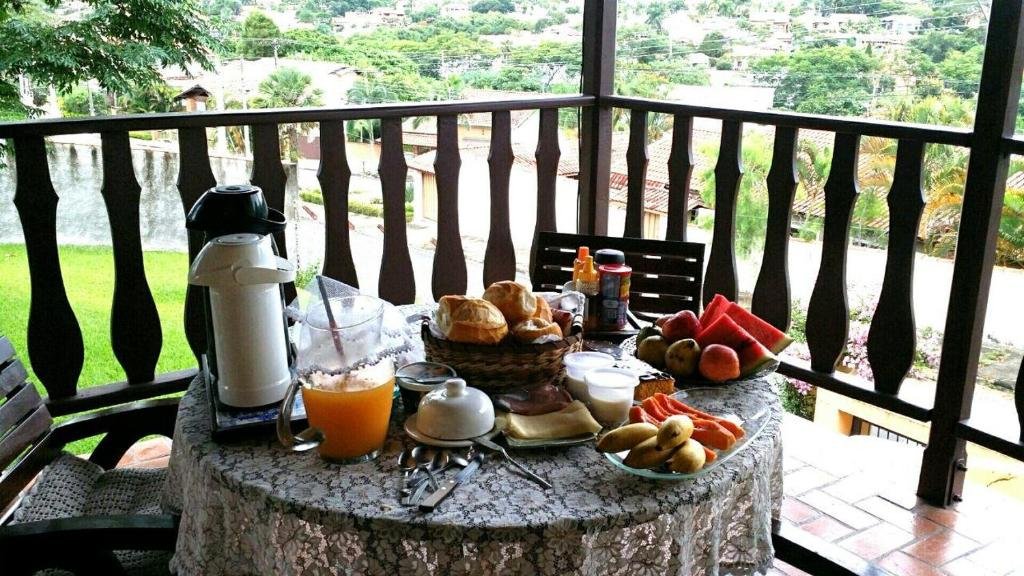 Uma mesa redonda com vários alimentos de café da manhã na Pousada Suítes Águas de São Pedro. A mesa está situada em uma varanda, e possui suco, café, pães, frios e frutas. 