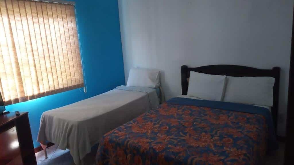 Quarto do airbnb Cantinho da Serra. No lado direito da imagem há uma cama de casal, já no lado esquerdo há uma cama de solteiro e ainda no lado esquerdo há uma cortina com persianas e é possível ver apenas um pedaço de uma cômoda. Imagem para ilustrar o post airbnb em Itatiaia.