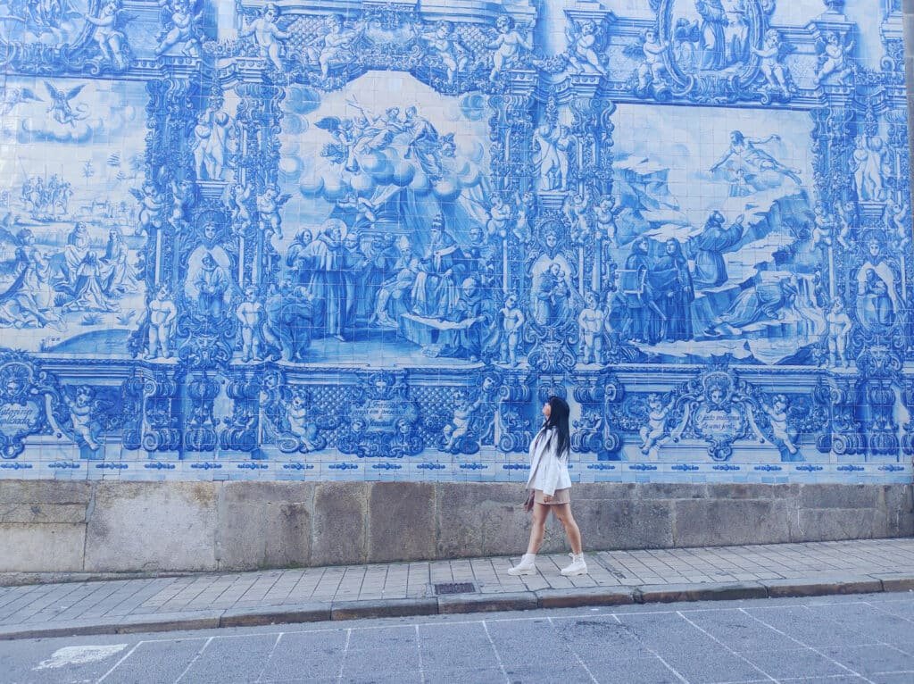 Uma pessoa vestida de branco está em frente a um grande mural azul e branco em uma parede de azulejos azuis, a Capela das Almas, em Porto, Portugal, olhando para a obra de arte. Esse é um dos destinos indicados no guia de mochilão pela Europa.