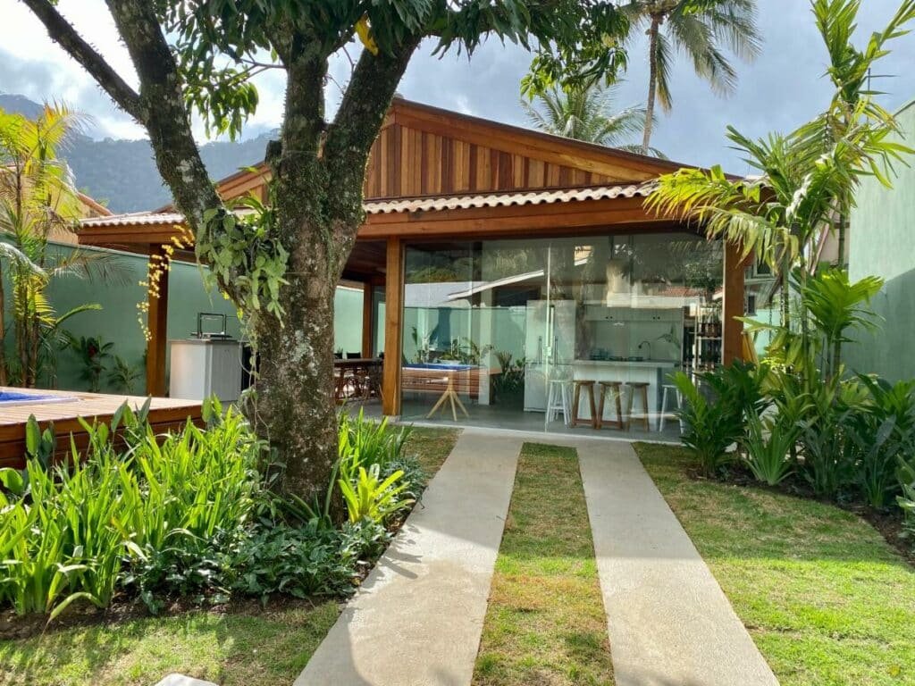 Frente da Casa Amendoeiras durante o dia com jardim a frente, uma árvore do lado esquerdo da imagem e ao fundo a propriedade.