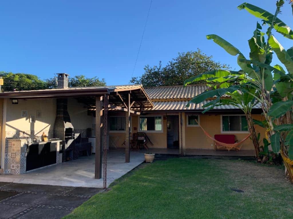 Área externa do airbnb Casa de Férias. No lado direito da imagem há algumas palmeiras e um gramado. No lado esquerdo há uma área coberta com pia, armários, churrasqueira, mesas e cadeiras e no fundo da imagem está a casa.