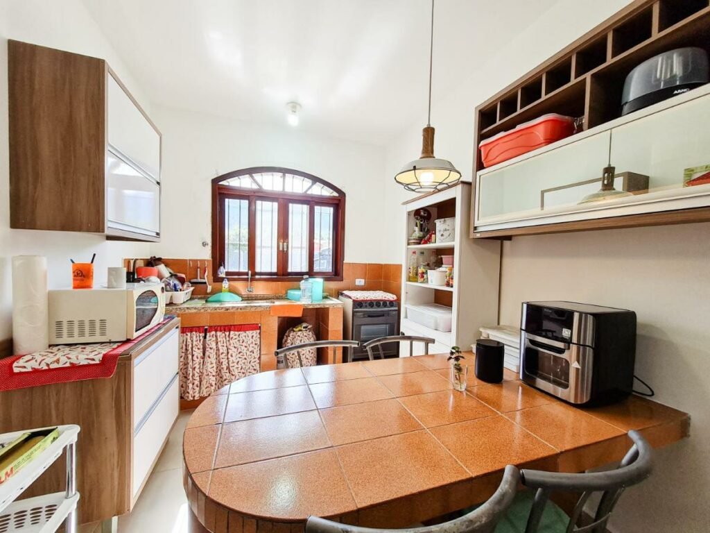 Cozinha bem iluminada da Casa vista para montanha Pet Friendly. Uma mesa com quatro cadeiras está em primeiro plano, e ao fundo pode-se ver as bancadas com utensílios, o micro-ondas e o fogão. Há uma janela em cima da pia.