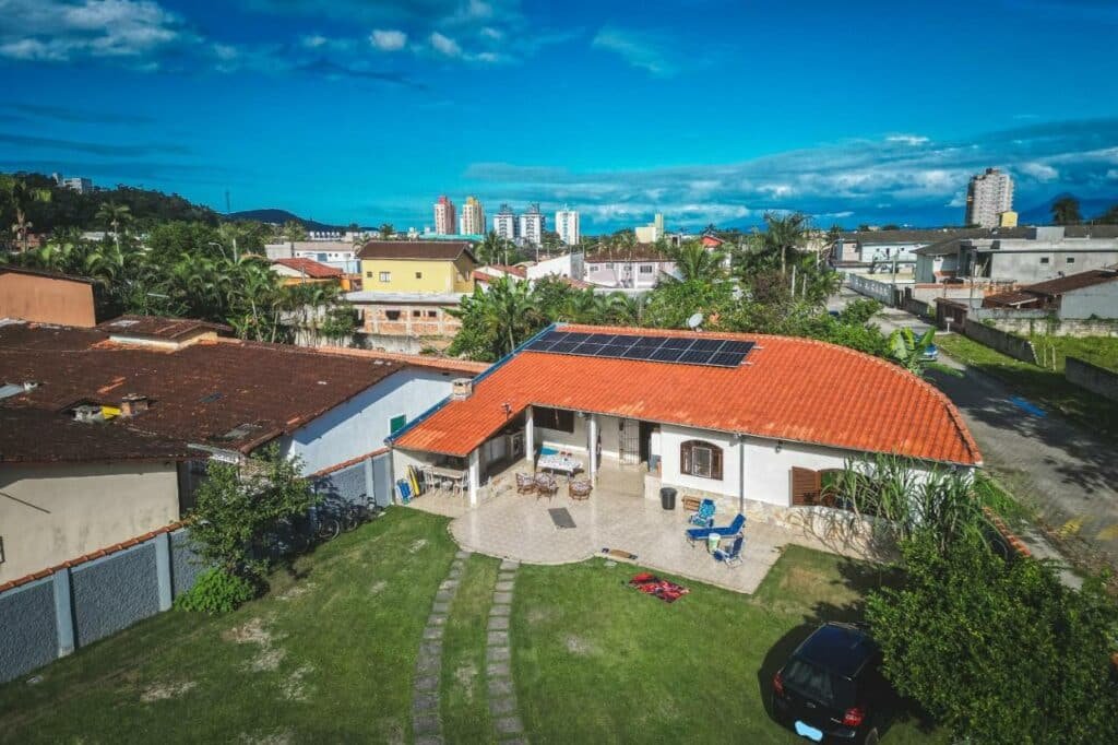 Vista aérea da Casa vista para montanha Pet Friendly. A propriedade tem um grande gramado em frente, e há um carro estacionado perto de uma árvore no canto inferior direito. O alpendre da casa também é espaçoso e conta com móveis de jardim. Ao fundo é possível ver prédios e casas da cidade.