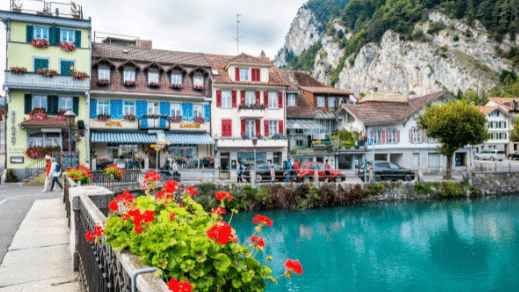 Imagem da cidade de Grindelwald com edifícios coloridos se alinham na orla de uma pequena cidade, com flores vermelhas em primeiro plano e montanhas ao fundo.