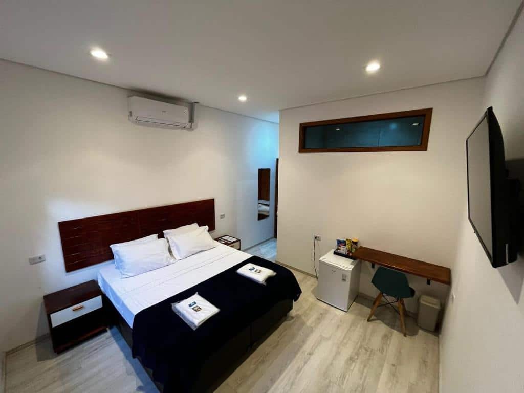 Uma cama no canto esquerdo com um ar-condicionado em cima. do lado o corredor da entrada e banheiro. No canto direito uma televisão, e atrás uma mesa de escritório com um frigobar.