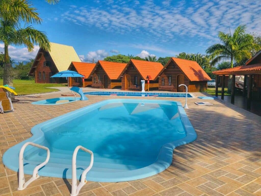 Duas piscinas com decorações aquáticas no Chalé Caiçara. Ao fundo há cinco chalés de madeira em estilo alpino. Uma faixa de grama pode ser vista na lateral esquerda, e há algumas palmeiras ao redor.