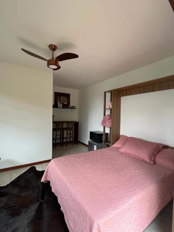 Quarto do airbnb Chale linda vista Penedo. No lado direito da imagem está a cama de casal, no lado esquerdo da cama há um frigobar e um microondas e no fundo da imagem há uma mesa com cadeiras e uma prateleira em cima. Imagem para ilustrar o post airbnb em Itatiaia.