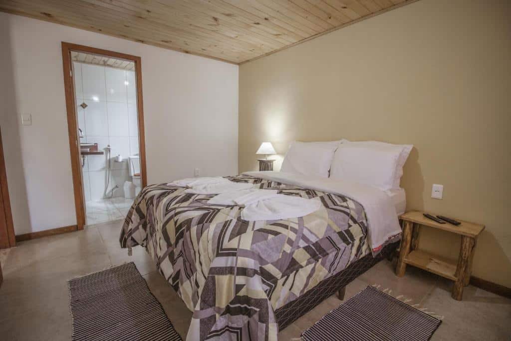 Imagem do quarto em Chalés Cerejeira, que está ilustrando o post sobre airbnb em Visconde de Mauá. É espaçoso e há uma cama box de casal no meio. Dos seus dois lados há uma mesa de cabeceira, e na da esquerda há um abajur. Há um banheiro na esquerda.