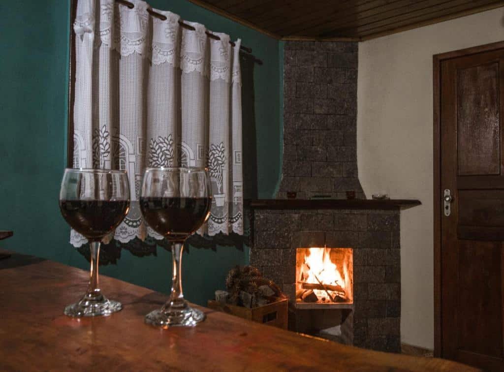 Sala no Chalés Cerejeira. Há uma bancada com duas taças de vinho perto da câmera, e no fundo há uma janela, lareira e uma porta.
