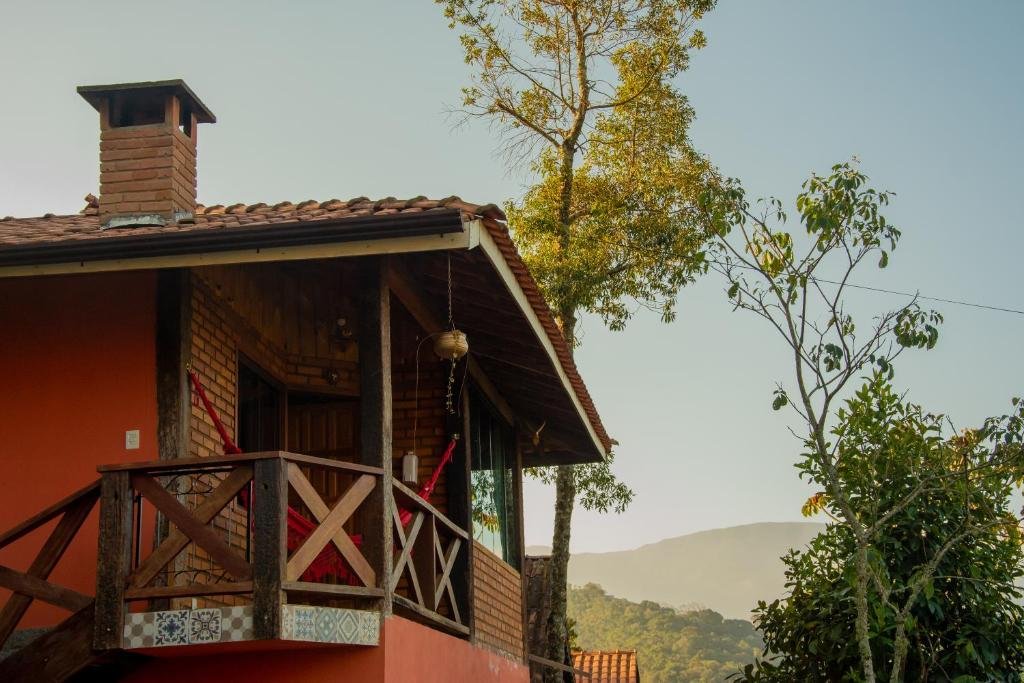 Exterior do Chalés Encanto do sol. A câmera aponta para o andar de cima, que conta com uma varanda. No fundo há árvores e montanhas.