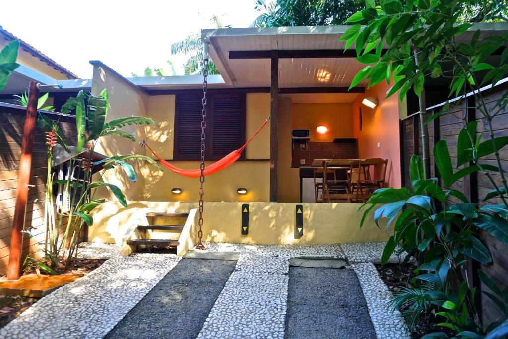 Entrada de um dos chalés Maritacas. Ele possui uma pequena escada de três degraus para entrar. Na varanda há uma rede, uma mesa e (mais ao fundo) uma pia, um freezer e um micro-ondas. O espaço é rodeado por algumas plantas.