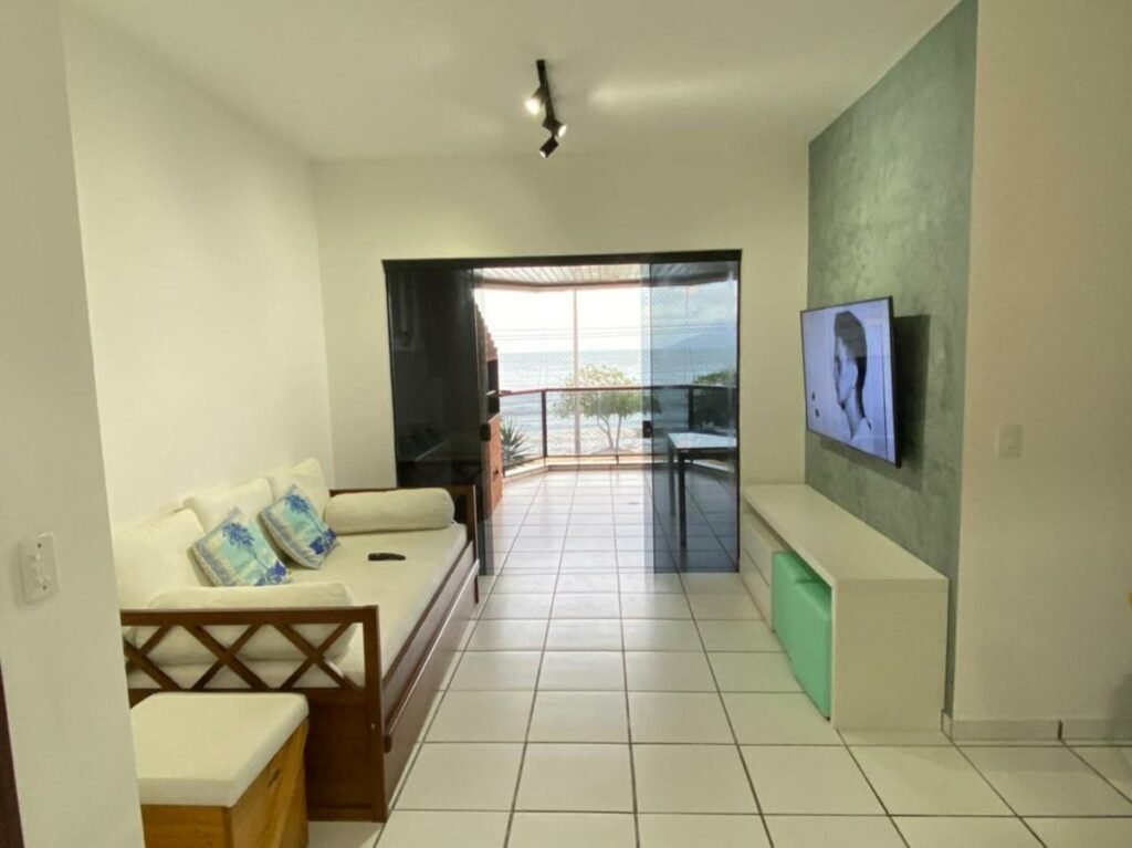 Sala de estar do Condomínio San Martim. Um sofá com almofadas está encostado na parede ao lado esquerdo e encara uma rack e uma TV logo em frente. Ao fundo do cômodo, ao lado esquerdo do sofá, está uma porta de vidro que leva à varanda com churrasqueira e vista da praia.