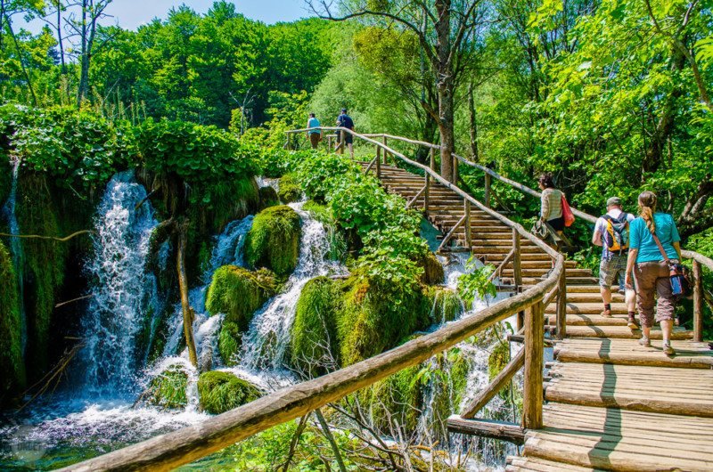 Pessoas caminhando sobre a ponte de madeira no Parque Nacional dos Lagos de Plitvice com vista para as cascatas.