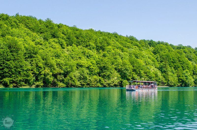 Um barco com viajantes flutuando sobre o lago do Parque Nacional dos Lagos de Plitvice. Há uma grande quantidade de árvores atrás.