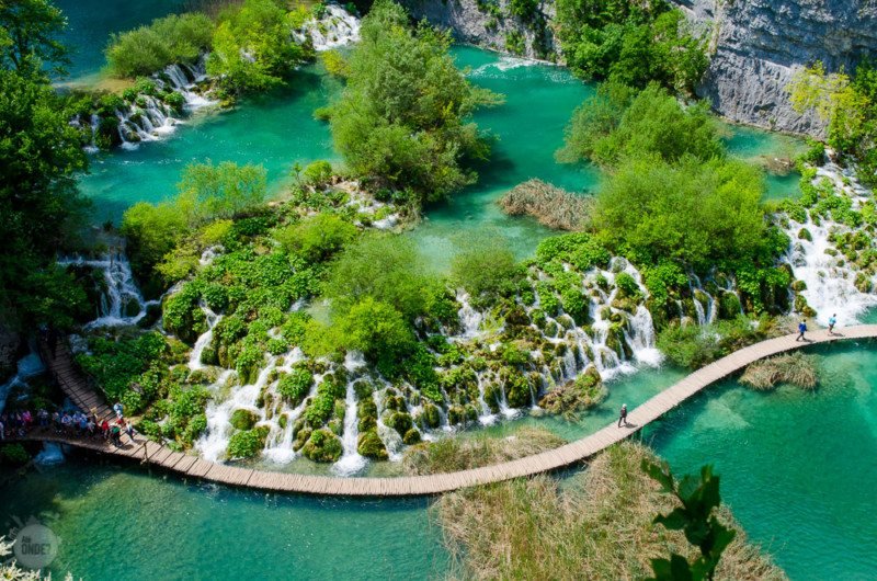 Foto aérea do Parque Nacional dos Lagos de Plitvice, mostrando o enorme lago que é cortado por árvores cachoeiras, montanhas uma ponte de madeira.