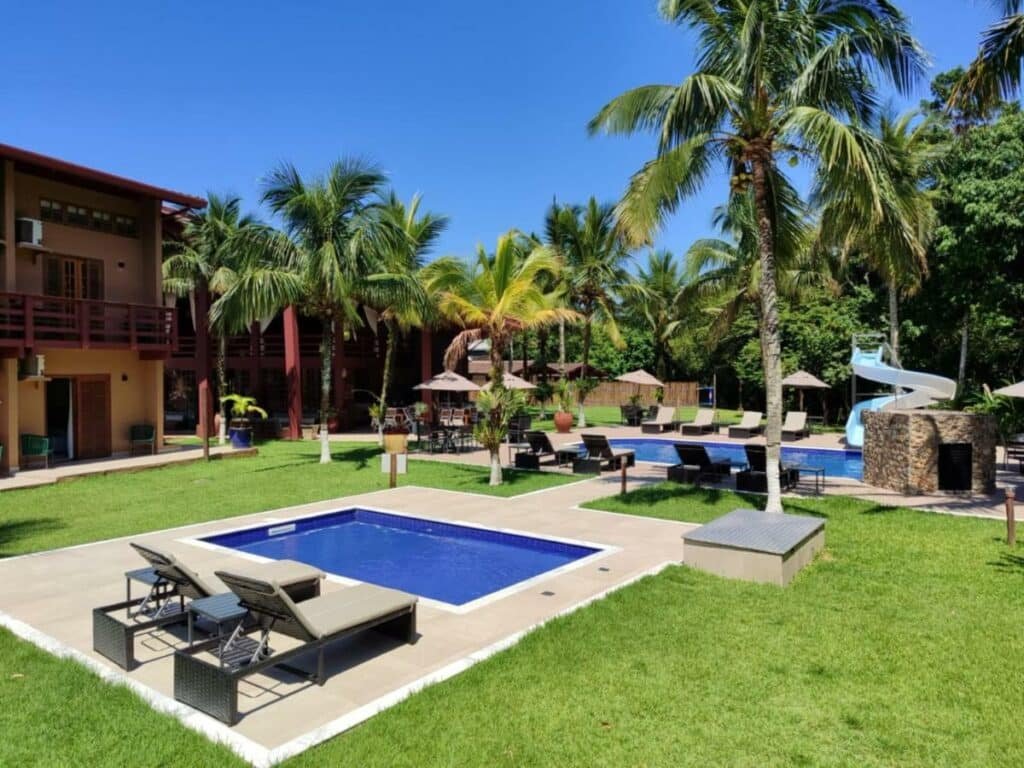 Piscinas da Delta Maresias Club by Smart Sharing. Ao fundo está a maior, com um toboágua e várias espreguiçadeiras ao redor. Mais em frente está a piscina menor e duas espreguiçadeiras. O local é aberto e tem várias árvores ao redor.