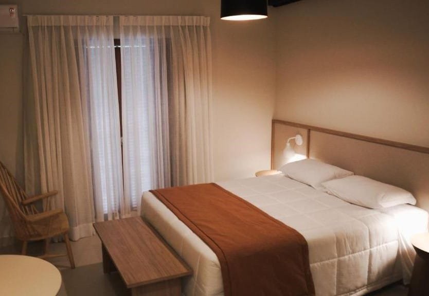 Quarto com uma cama de casal e um banquinho ao seus pés da Delta Maresias Club by Smart Sharing. Em frente a cama está uma cadeira e ao lado uma janela com cortina.