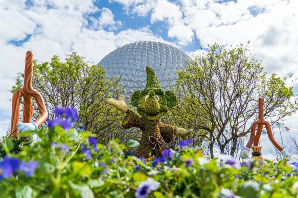 Epcot em Orlando com a famosa esfera de fundo com o Mickey de cartola de mágico esculpido em grama com flores violeta e árvores, o céu é azul com muitas nuvens