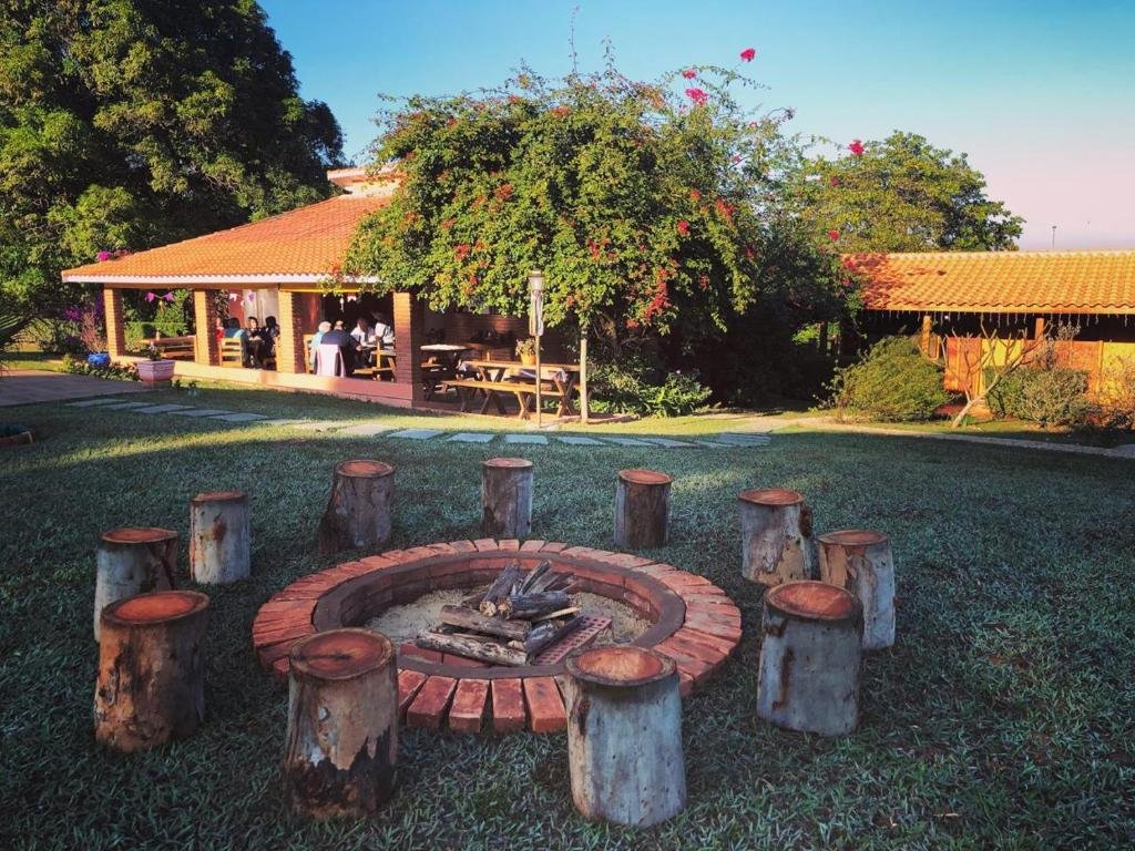 Área externa na Estância Travessia pousada. No centro da imagem vemos onze troncos de madeira cortados ao meio para servirem de bancos, e uma fogueira ao meio. Ao fundo, temos visão do rancho da pousada, onde há pessoas sentadas conversando.