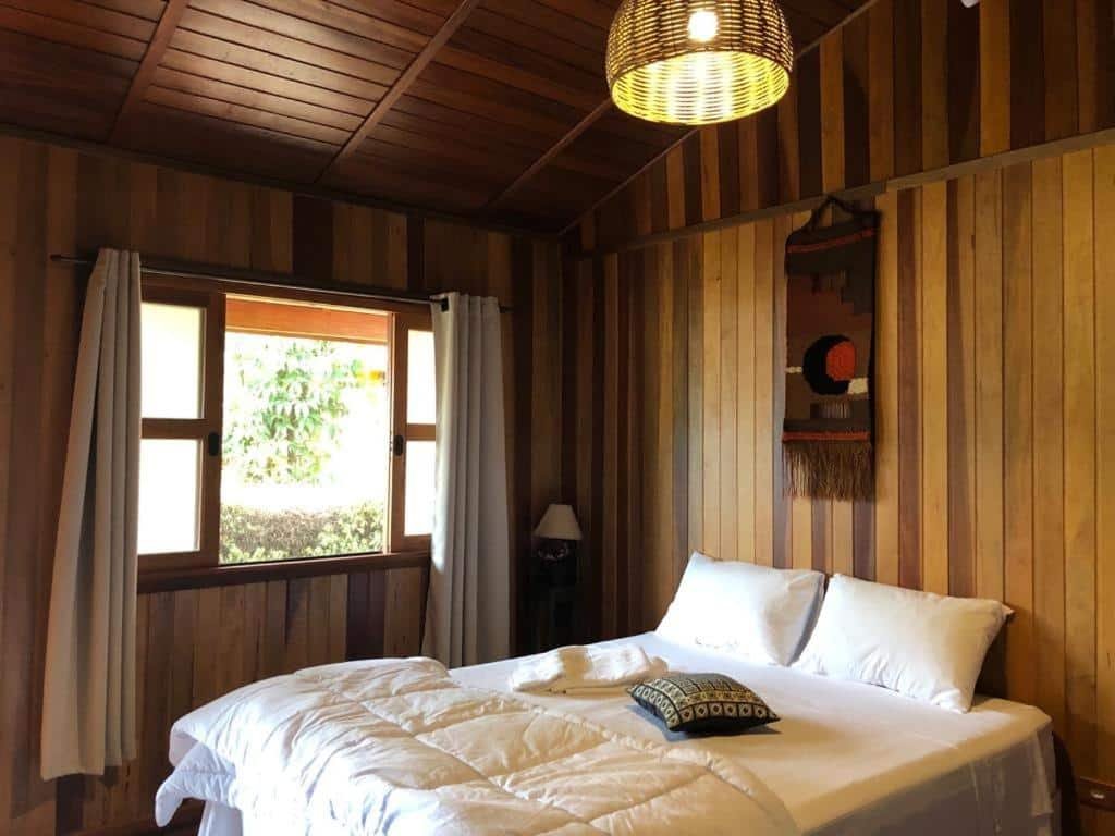 Um quarto da Estância Travessia, em São Pedro, para representar o post sobre pousadas em Águas de São Pedro. As paredes do quarto são de madeira, há uma janela com cortinas, um abajur no canto do Cômodo e uma cama de casal com lençóis brancos.