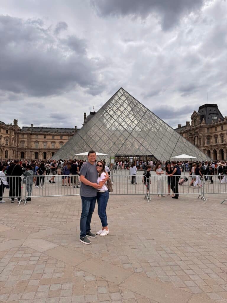 Um casal está em frente à entrada da pirâmide de vidro do Museu do Louvre, em Paris, cercado por outros visitantes. Esse é um dos destinos indicados no guia de mochilão pela Europa.