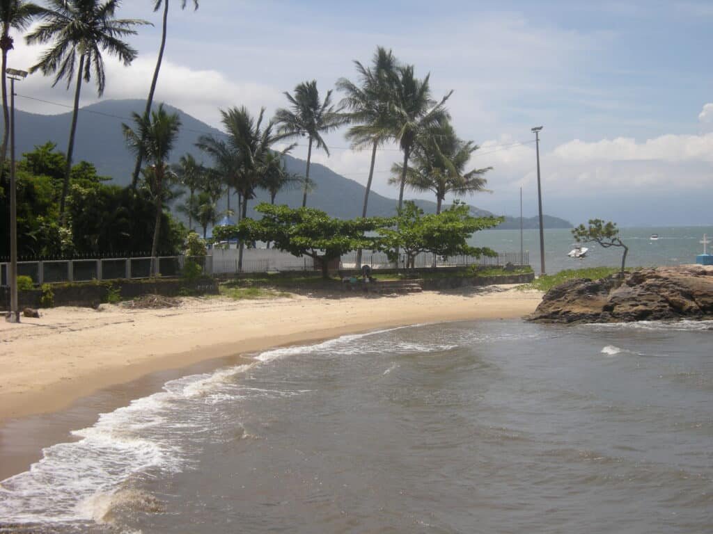 Praia do Pontal da Cruz com ondas suaves, palmeiras e uma montanha distante sob um céu parcialmente nublado. Cerca e barcos visíveis. No canto direito é possível ver uma cruz entre as rochas.
