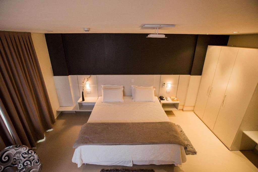 Quarto do Golden Plaza Hotel. No meio uma cama de casal, de cada lado uma prateleira e um abajur. Do lado esquerdo uma poltrona com cortina, do lado direito um guarda-roupa. Foto para ilustrar post sobre hotéis em Porto Velho.