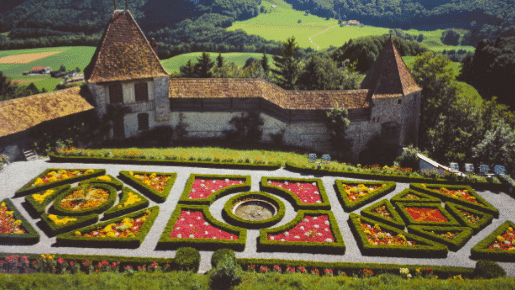Vista aérea de um jardim formal em Gruyères com padrões geométricos e diversas flores coloridas, cercado por árvores e muros de pedra.