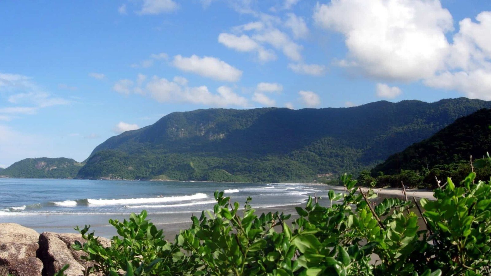 Vista panorâmica da praia de Guaecá com ondas suaves, vegetação verdejante em primeiro plano e montanhas arborizadas sob um céu azul.
