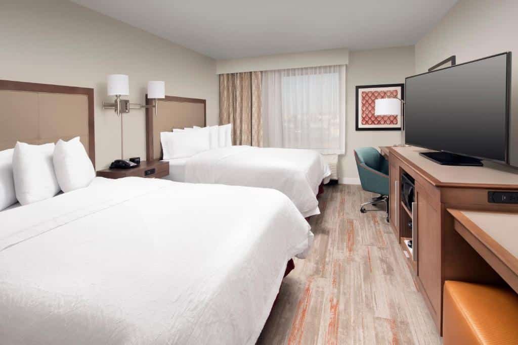 Quarto do Hampton Inn & Suites San Antonio Lackland AFB SeaWorld com duas camas de casal, uma janela com persianas, uma mesa de escritório com uma cadeira giratória, um móvel de madeira com gavetas e uma televisão sob ele, para representar onde ficar em San Antonio