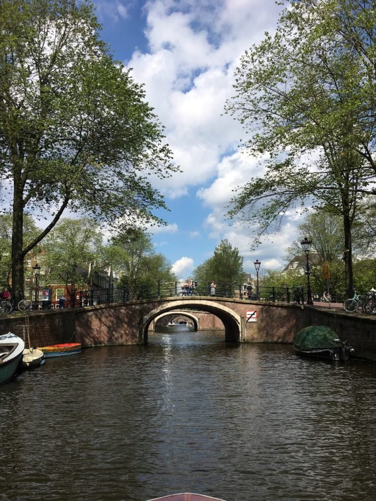 Vista do canal de Amsterdam com margens arborizadas, pequenos barcos e duas pontes de tijolos em arco sob um céu azul com nuvens fofas. Esse é um dos destinos indicados no guia de mochilão pela Europa.