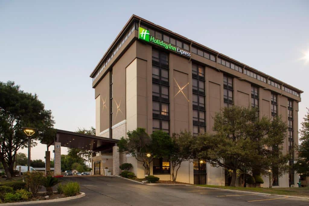 Prédio do Holiday Inn Express - San Antonio Airport, an IHG Hotel com oito andares, pintado em tons de bege e marrom, há algumas árvores nos arredores e vagas para estacionar